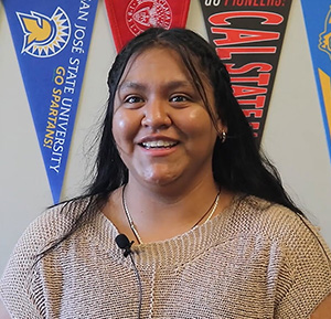 Student smiling at camera, with a variety of college banners behind her